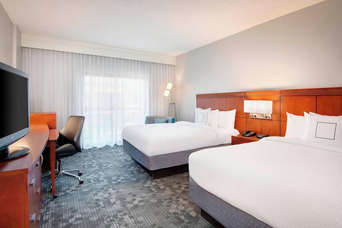 A hotel room with two beds, a desk with a TV, a chair, a lamp, and a window with curtains.
