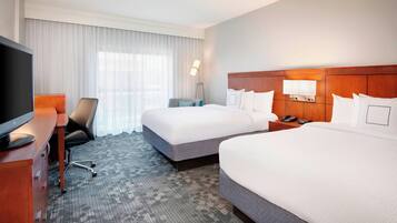 A hotel room with two beds, a desk with a TV, a chair, a lamp, and a window with curtains.