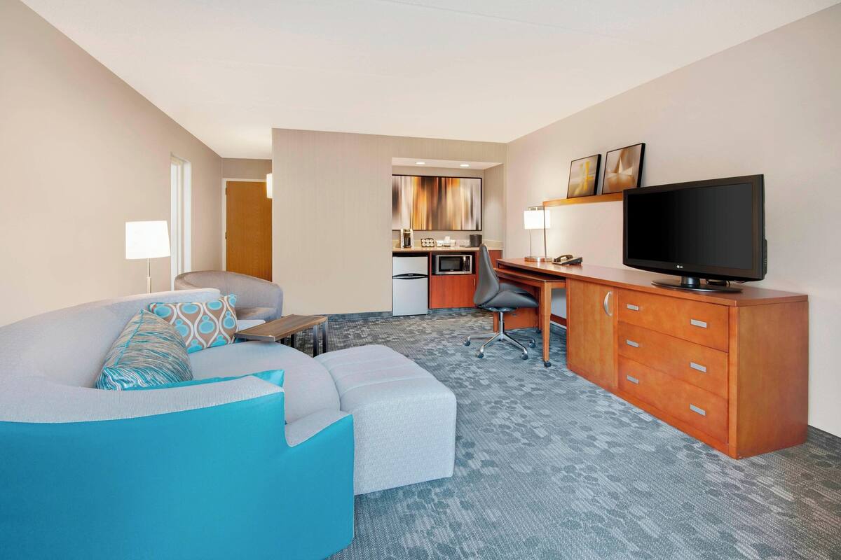 A hotel room with a large sofa, a desk with a computer, a television, and a kitchenette in the background.