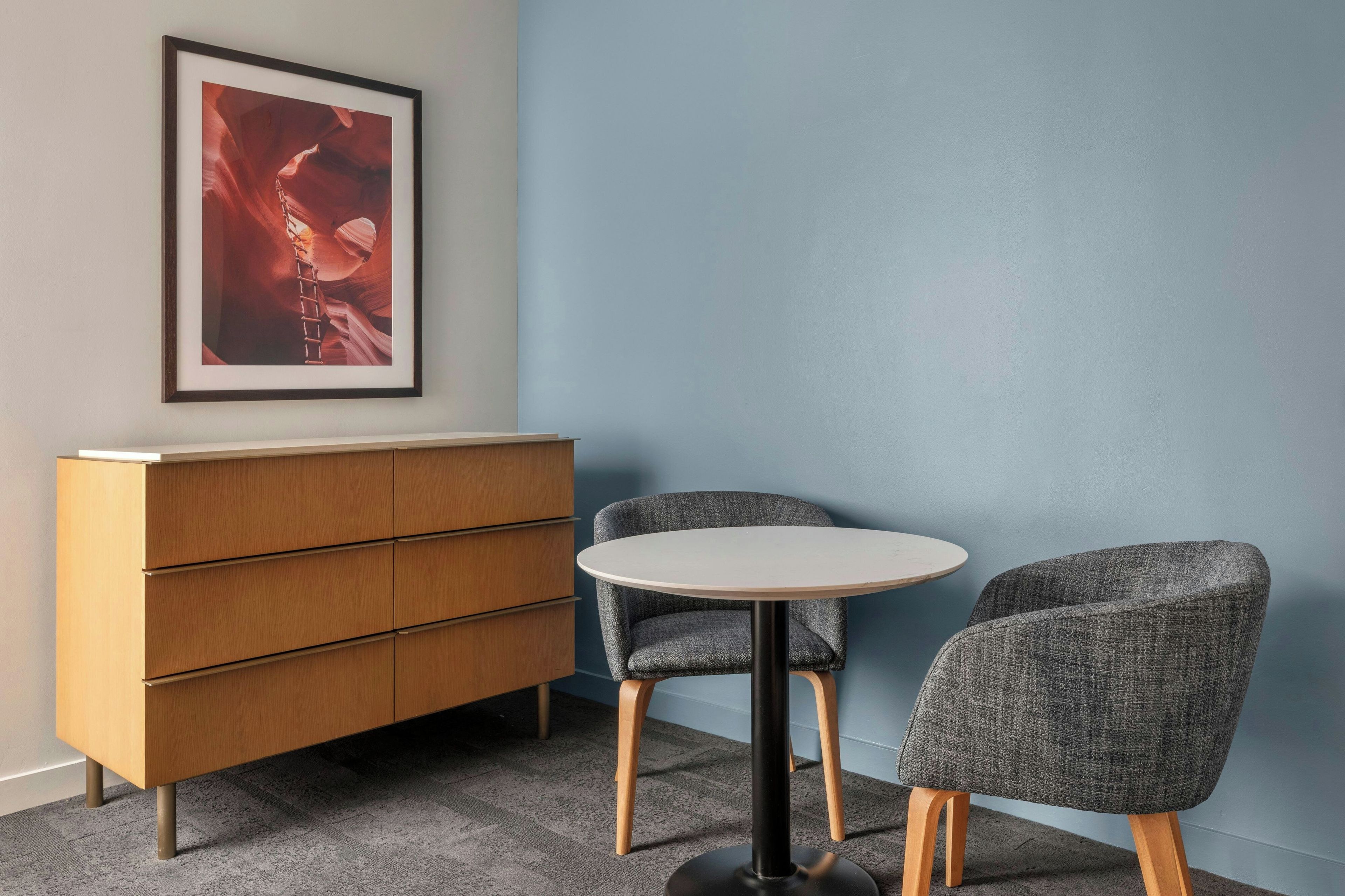 Une pièce comprenant un meuble en bois, deux fauteuils gris à revêtement en tissu, une table ronde et un tableau encadré accroché au mur.