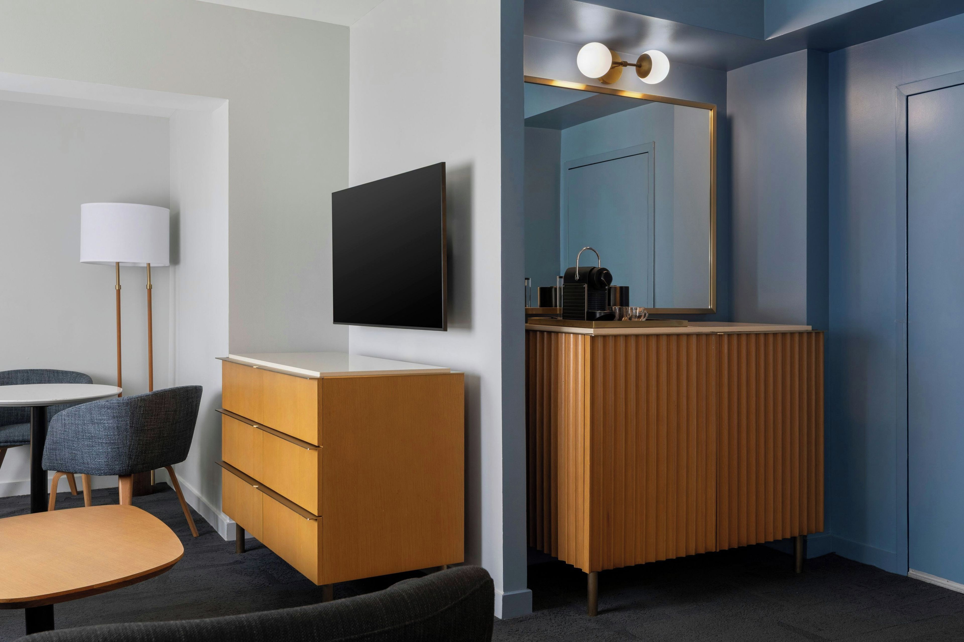 A hotel room with a flat-screen TV mounted on the wall, a wooden vanity with a mirror, a round table with two chairs, and a lamp.