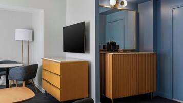 A hotel room with a flat-screen TV mounted on the wall, a wooden vanity with a mirror, a round table with two chairs, and a lamp.