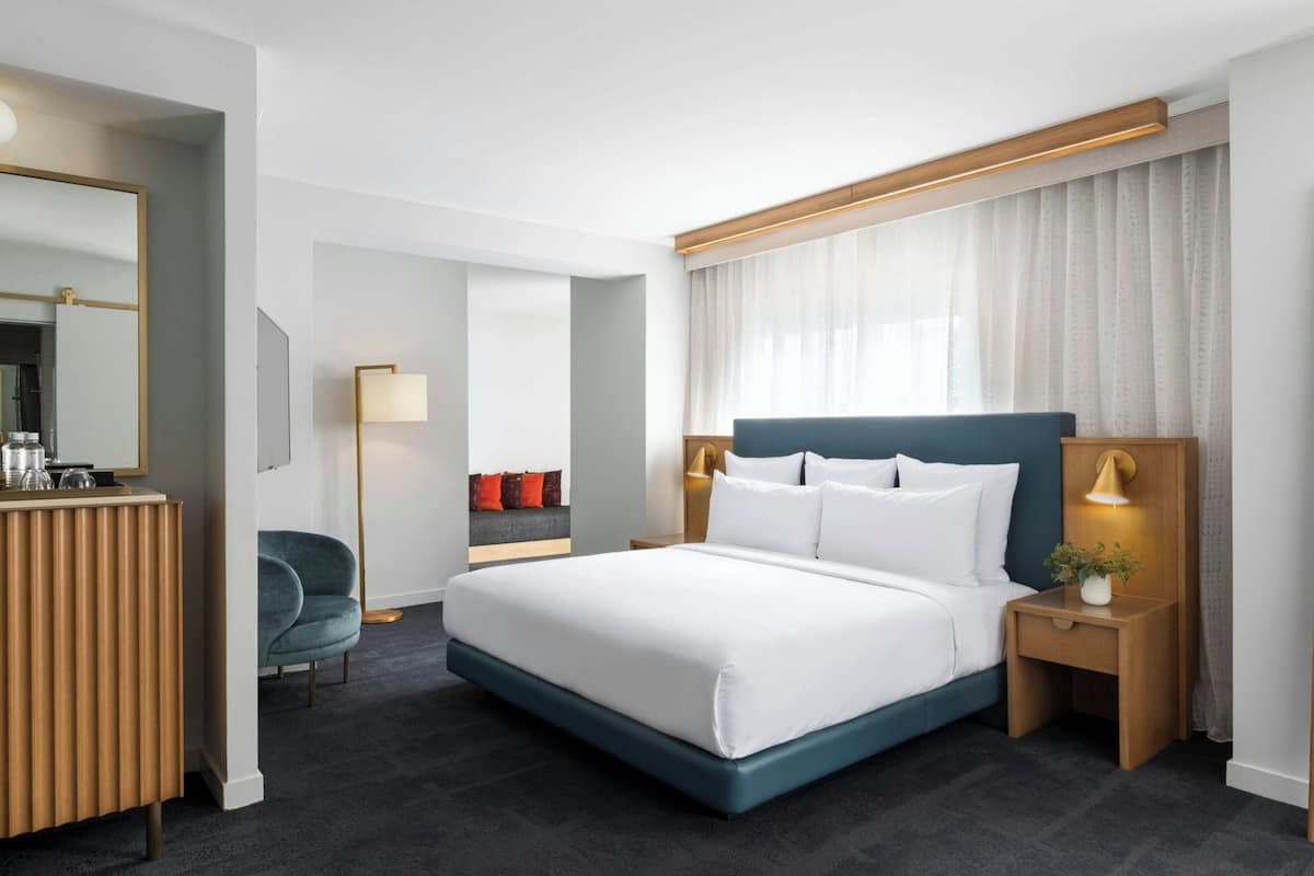 A hotel room with a large bed, bedside tables, a desk with a mirror, a chair, and a view of the outside through a window with curtains.