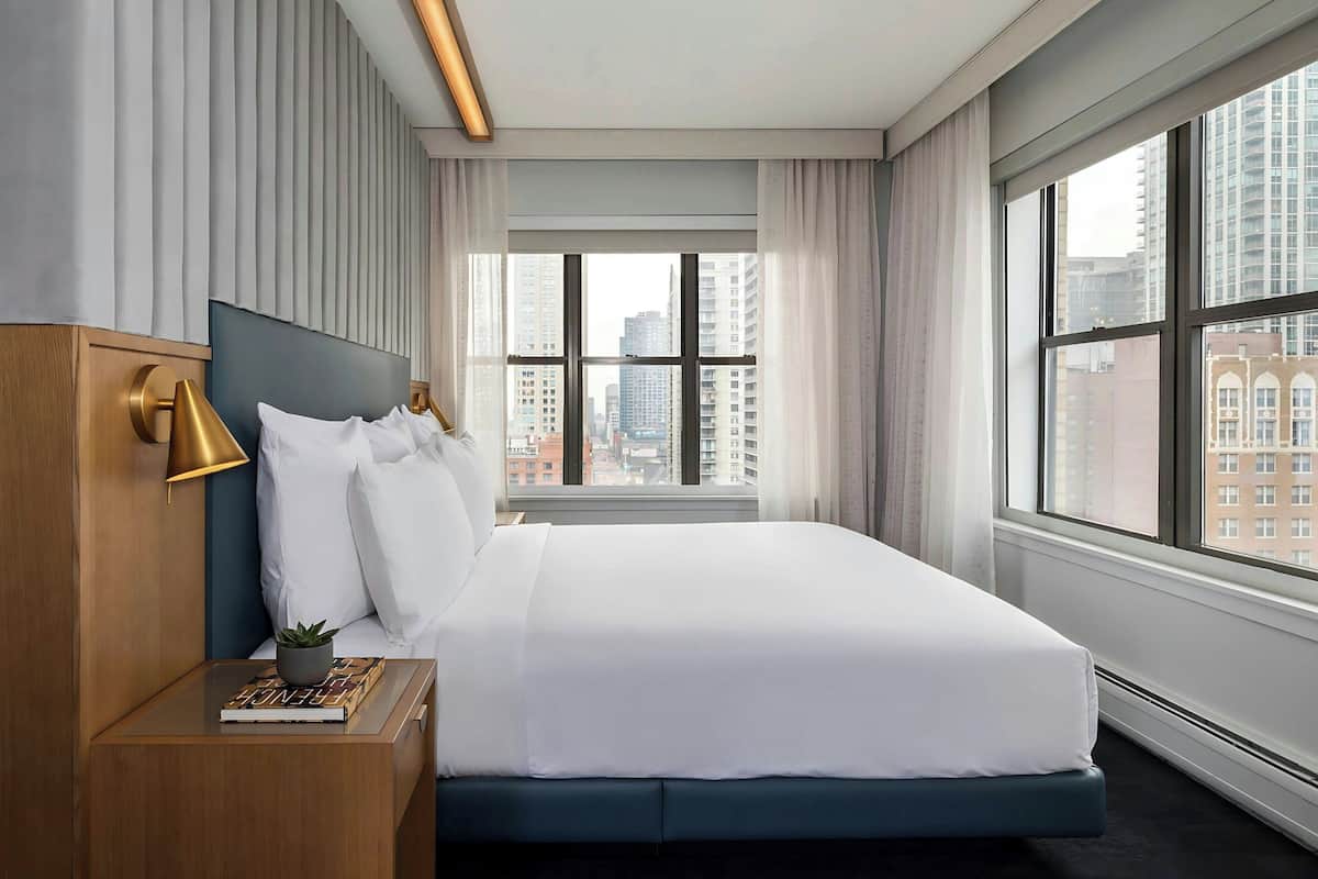 A modern hotel room with a large bed, a bedside table with a lamp, a view of the city through large windows, and a cityscape visible outside.
