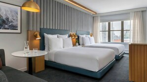 A modern hotel room with two beds, a small table with glasses, a wall-mounted lamp, and a city view.