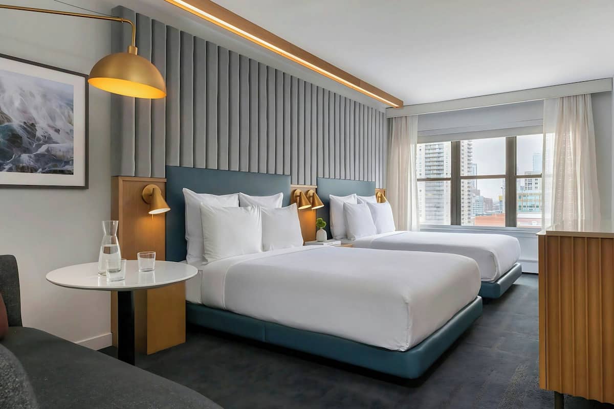 A modern hotel room with two beds, a small table with glasses, a wall-mounted lamp, and a city view.