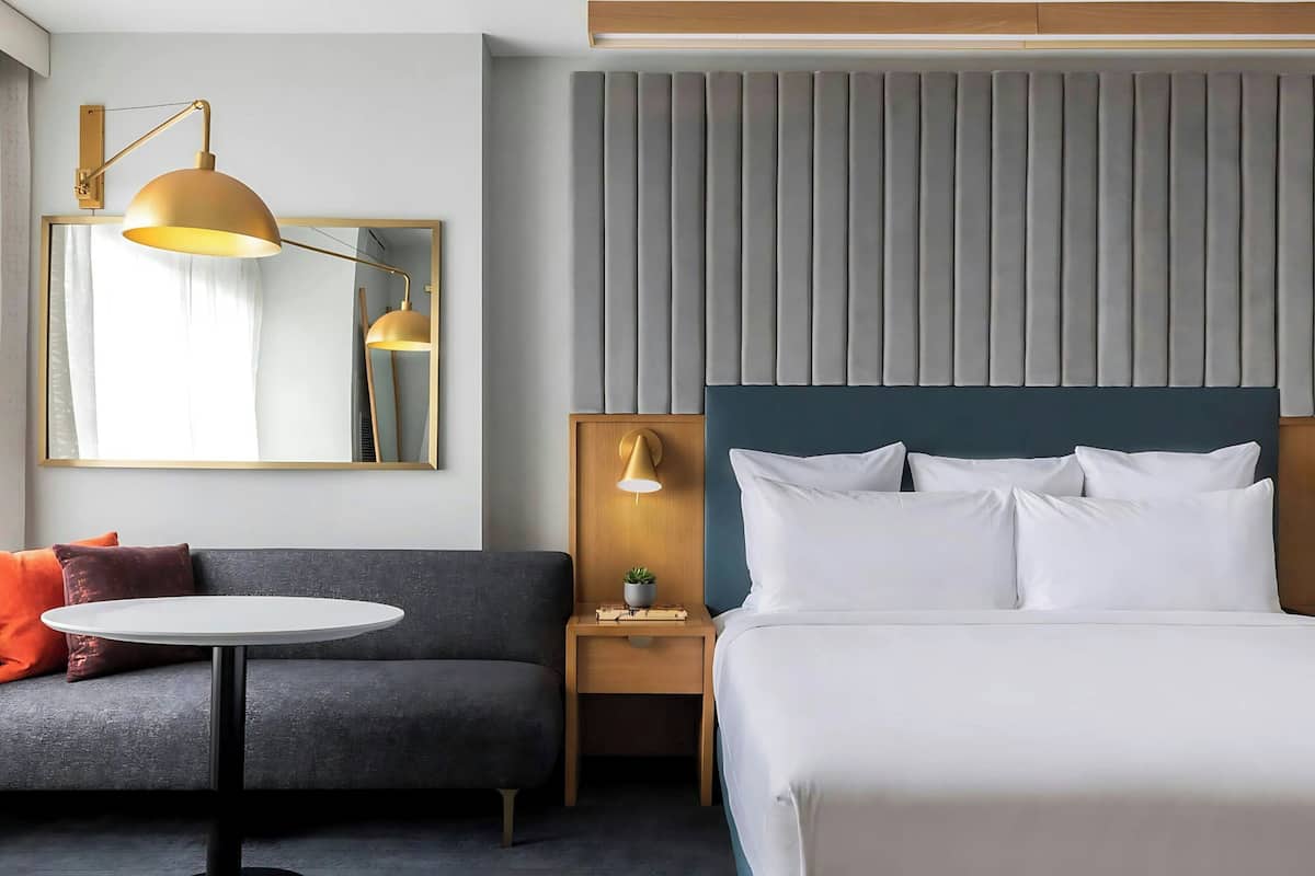 A hotel room with a bed, a bedside table with a lamp, a round table, a sofa, and a large mirror.