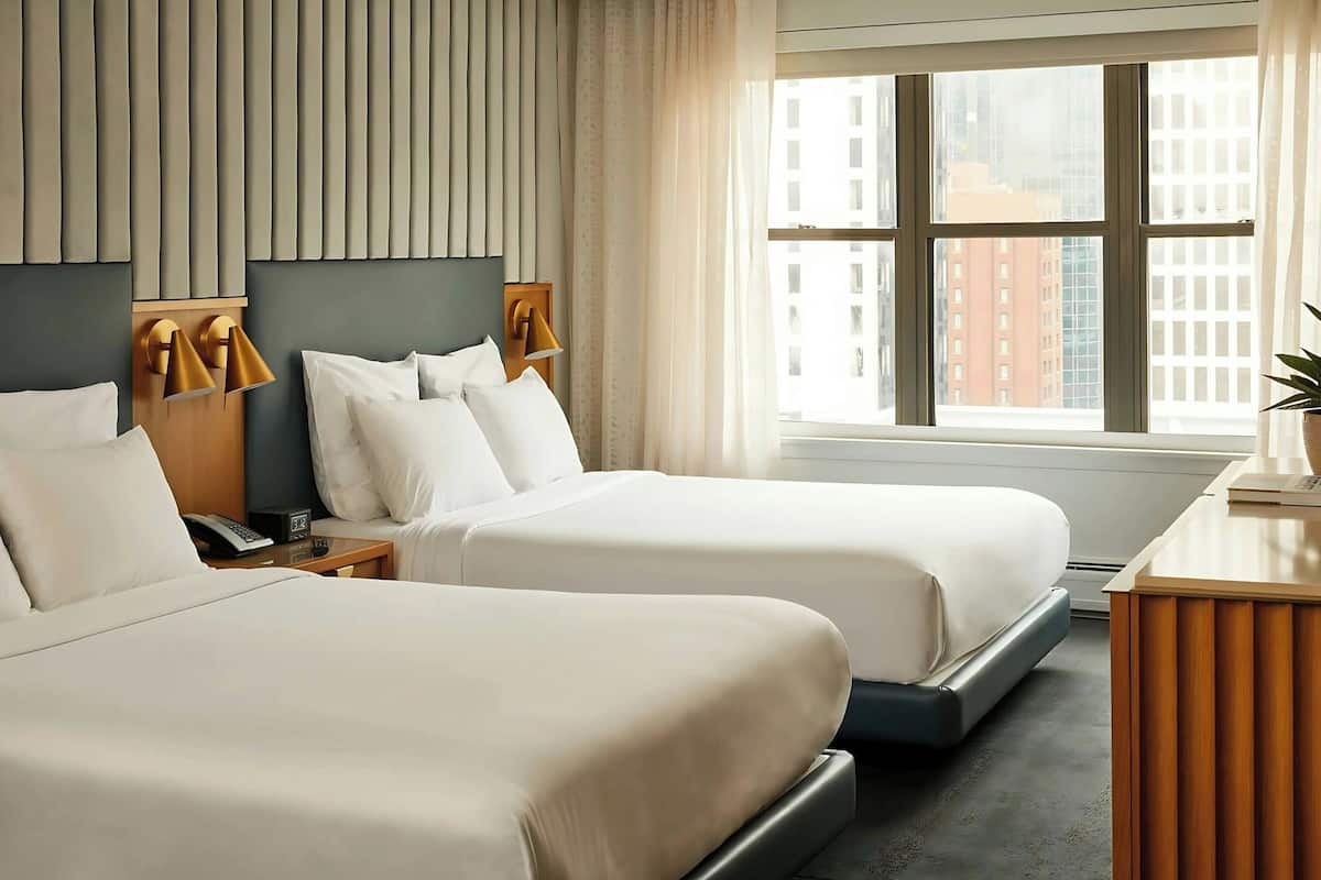 A hotel room with two beds, a nightstand with a phone, a desk with a lamp, and a window with a city view.