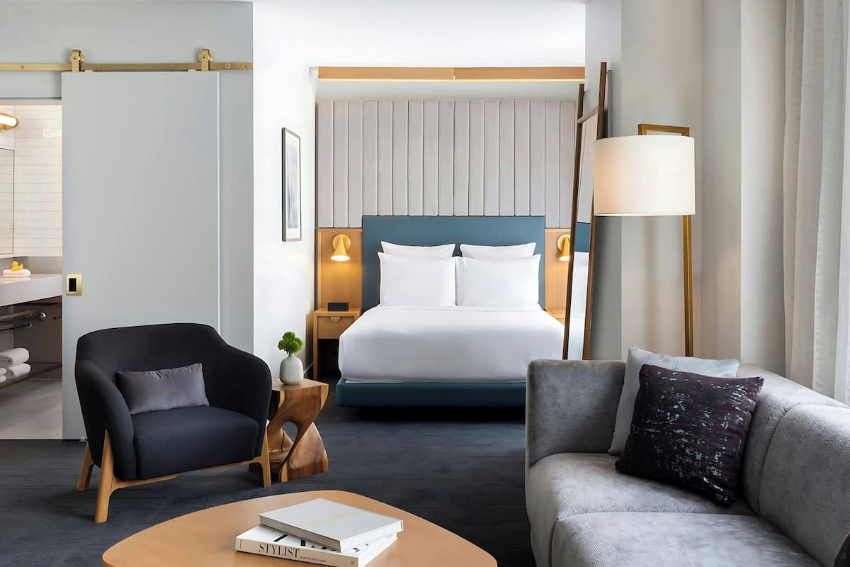 A hotel room with a bed, two chairs, a bedside table, a lamp, and a small plant.