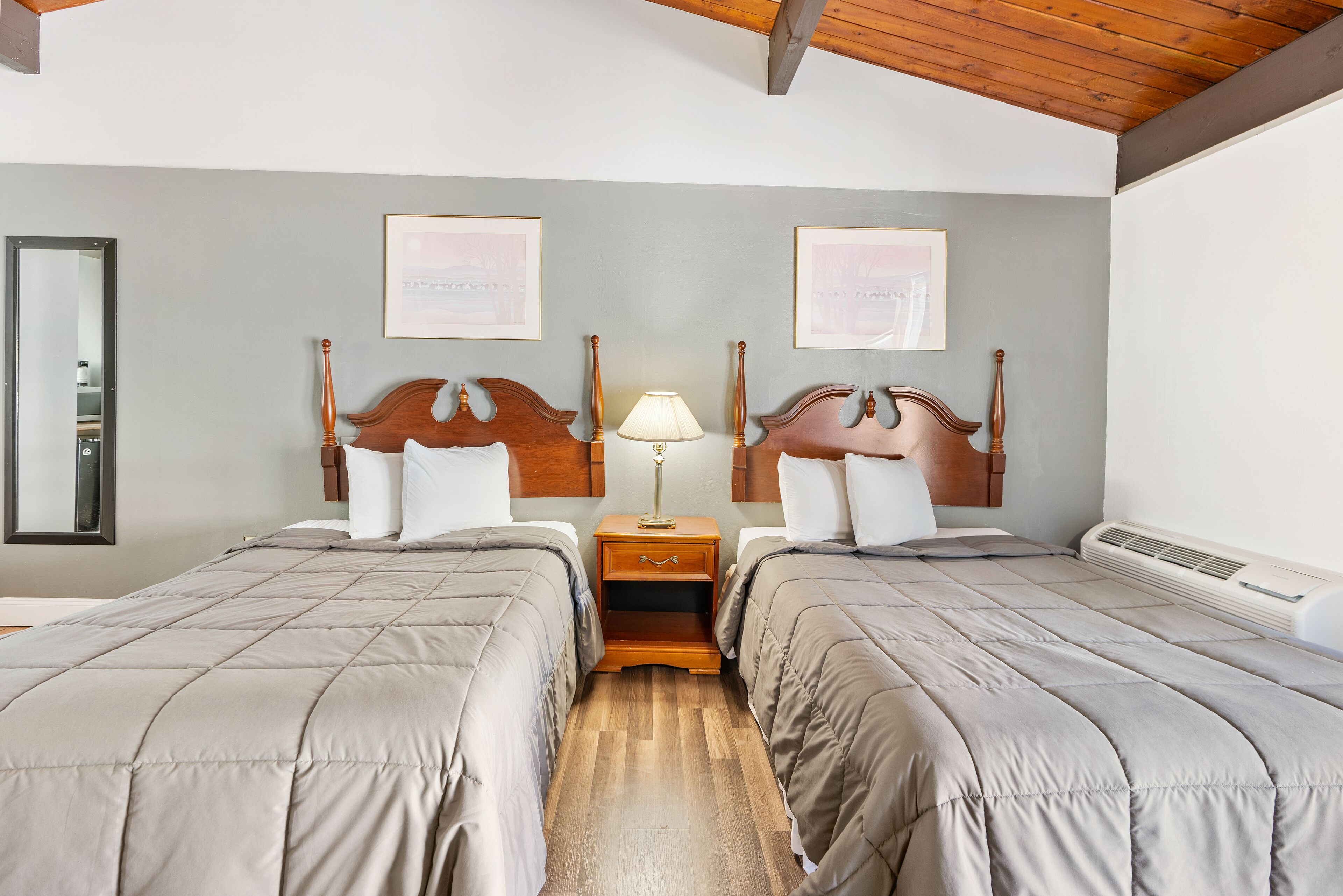 A hotel room with two beds, wooden headboards, a nightstand with a lamp, and framed pictures on the wall.