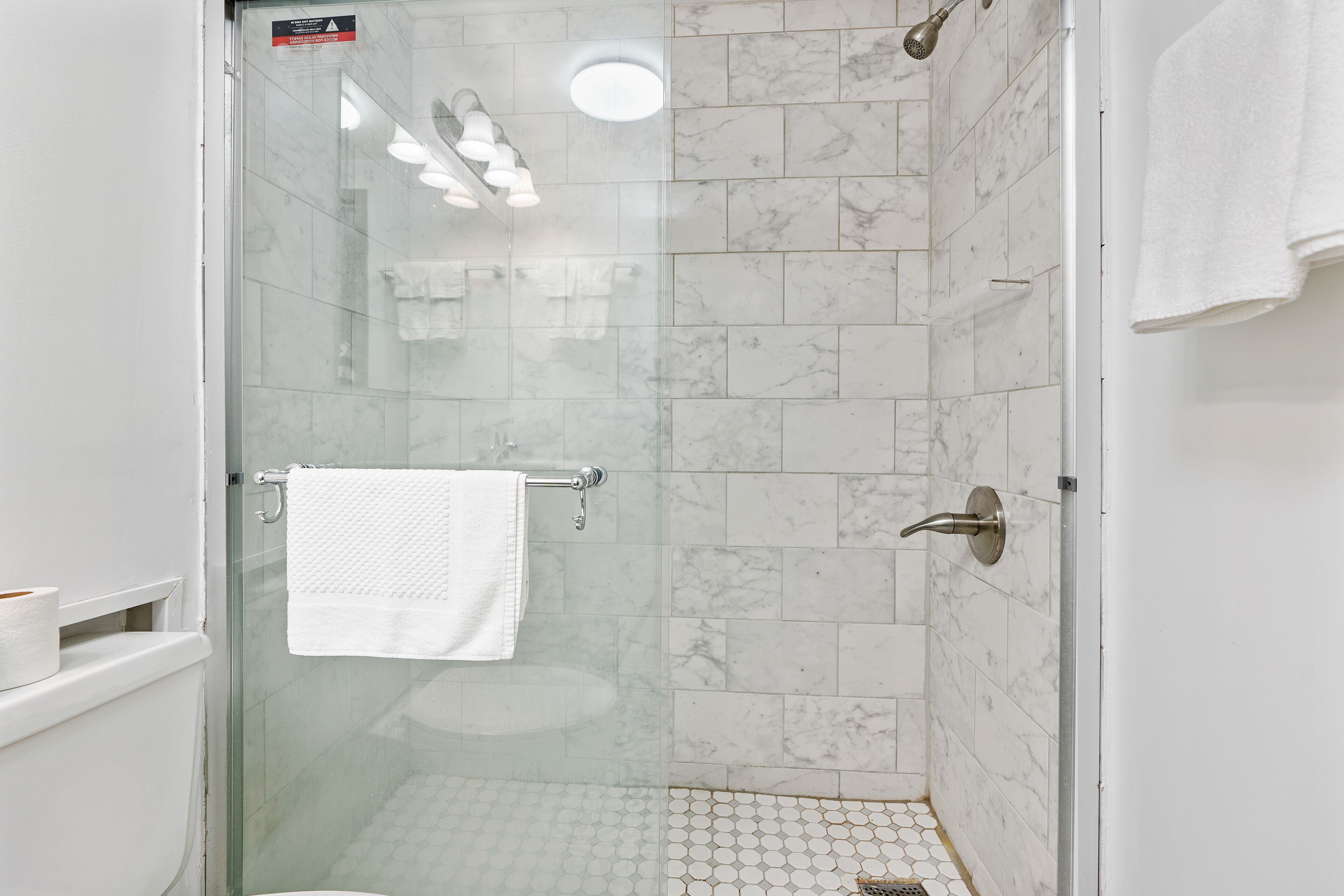 A glass-enclosed shower with white marble tiles, a glass door, and a white towel hanging on a metal towel rack.