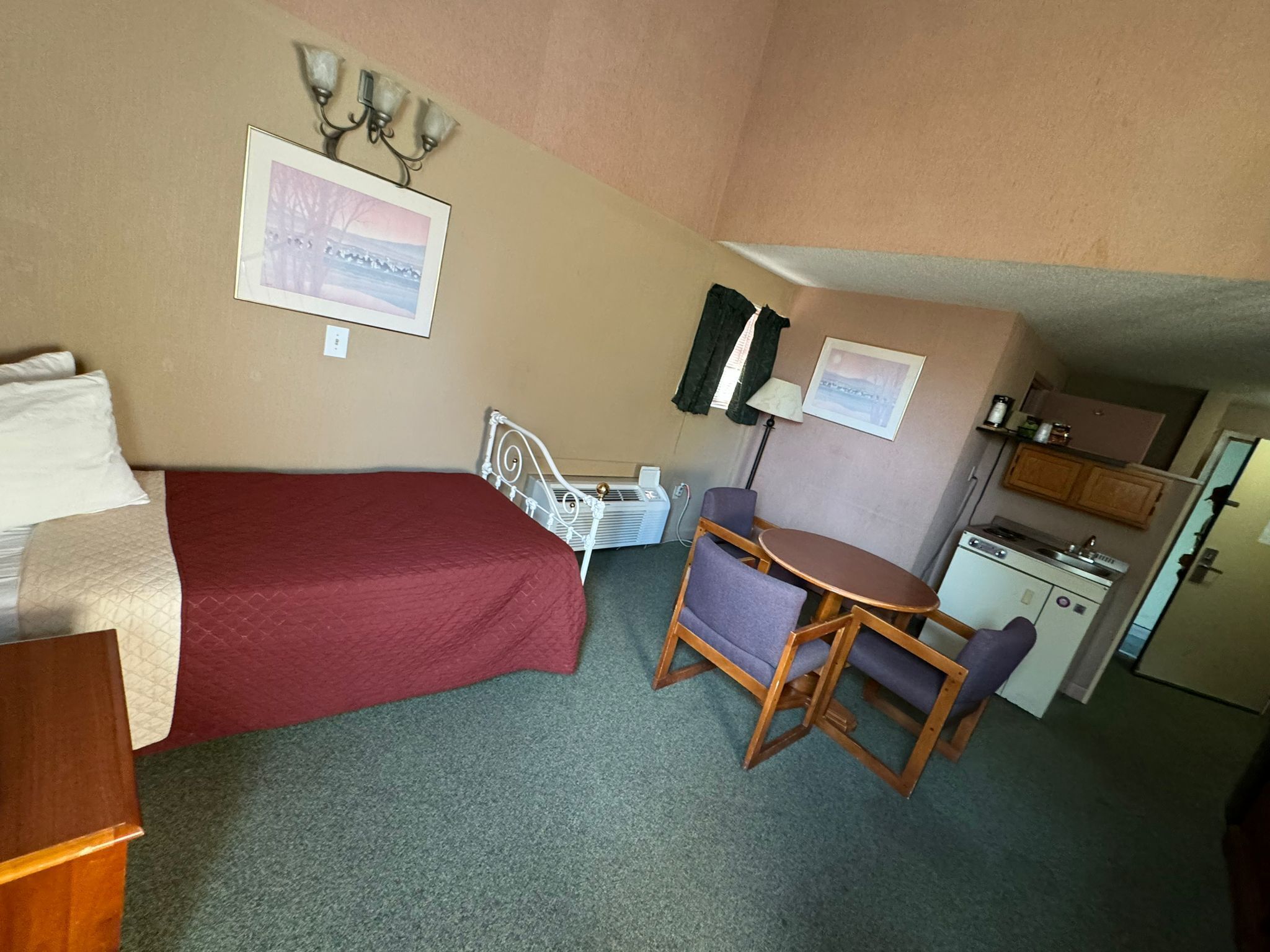 A small, compact room with a single bed, a desk, a chair, a kitchenette, and a small table.