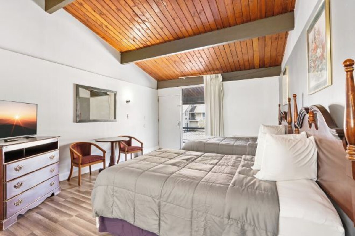 Une chambre avec un plafond en bois, un lit, un bureau avec une chaise, une télévision et une fenêtre avec des rideaux.