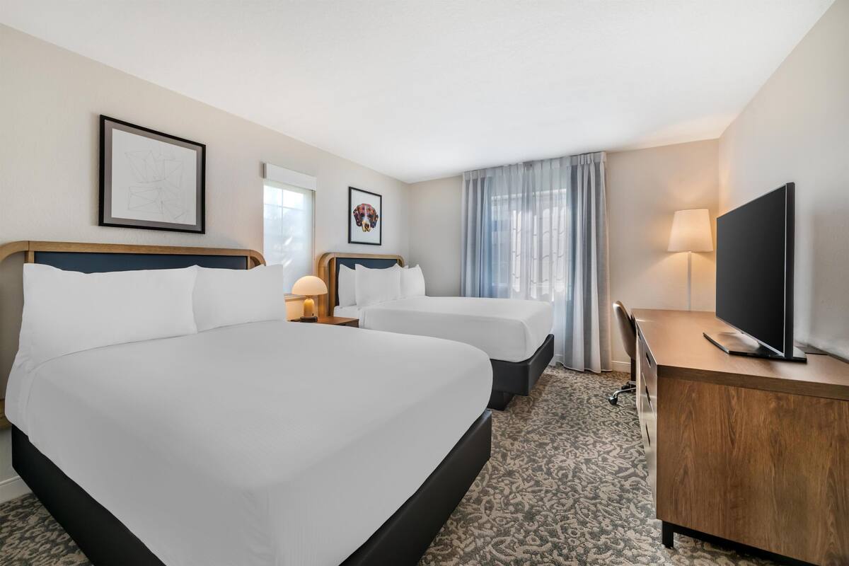 Une chambre d’hôtel avec deux lits, un bureau, une télévision et une fenêtre avec des rideaux.