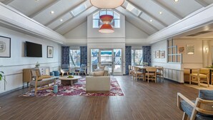 A spacious living area with a high ceiling, a large sofa, a dining table, and a TV mounted on the wall.