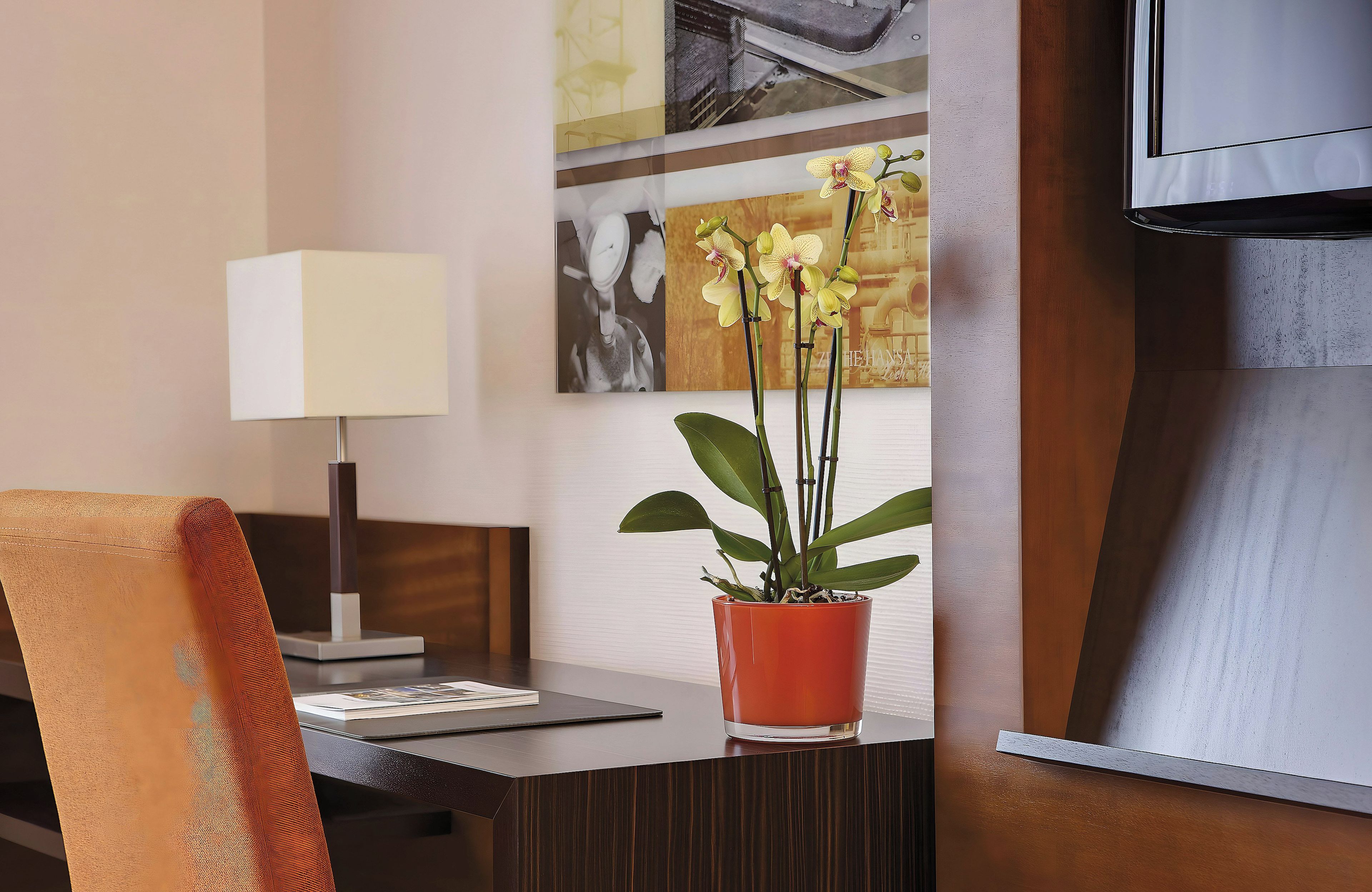 A hotel room with a desk, a lamp, a potted orchid, and a television.