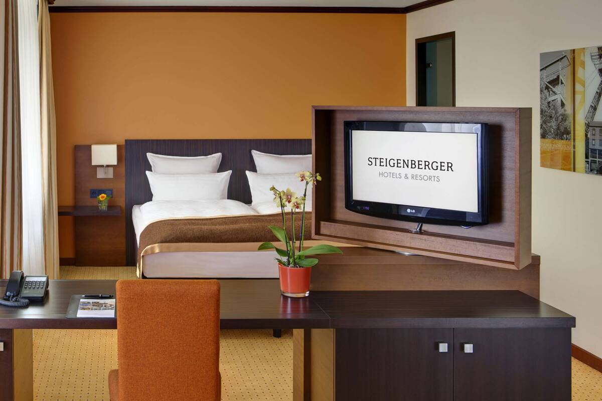 A hotel room with a bed, a desk with a telephone, a TV mounted on the wall, and a potted plant.