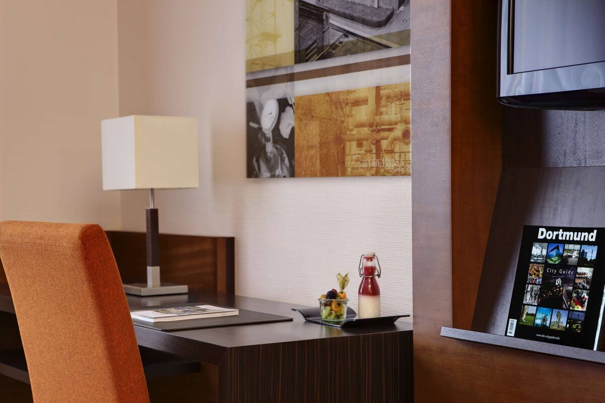 A hotel room with a wooden desk, a modern lamp, a city guide poster, and a small fruit bowl with a drink.