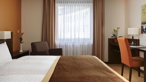 A hotel room with a large bed, a desk with a chair, a small table with a lamp, and a window with curtains. - Leonardo Royal Hotel Dortmund (Dortmund)