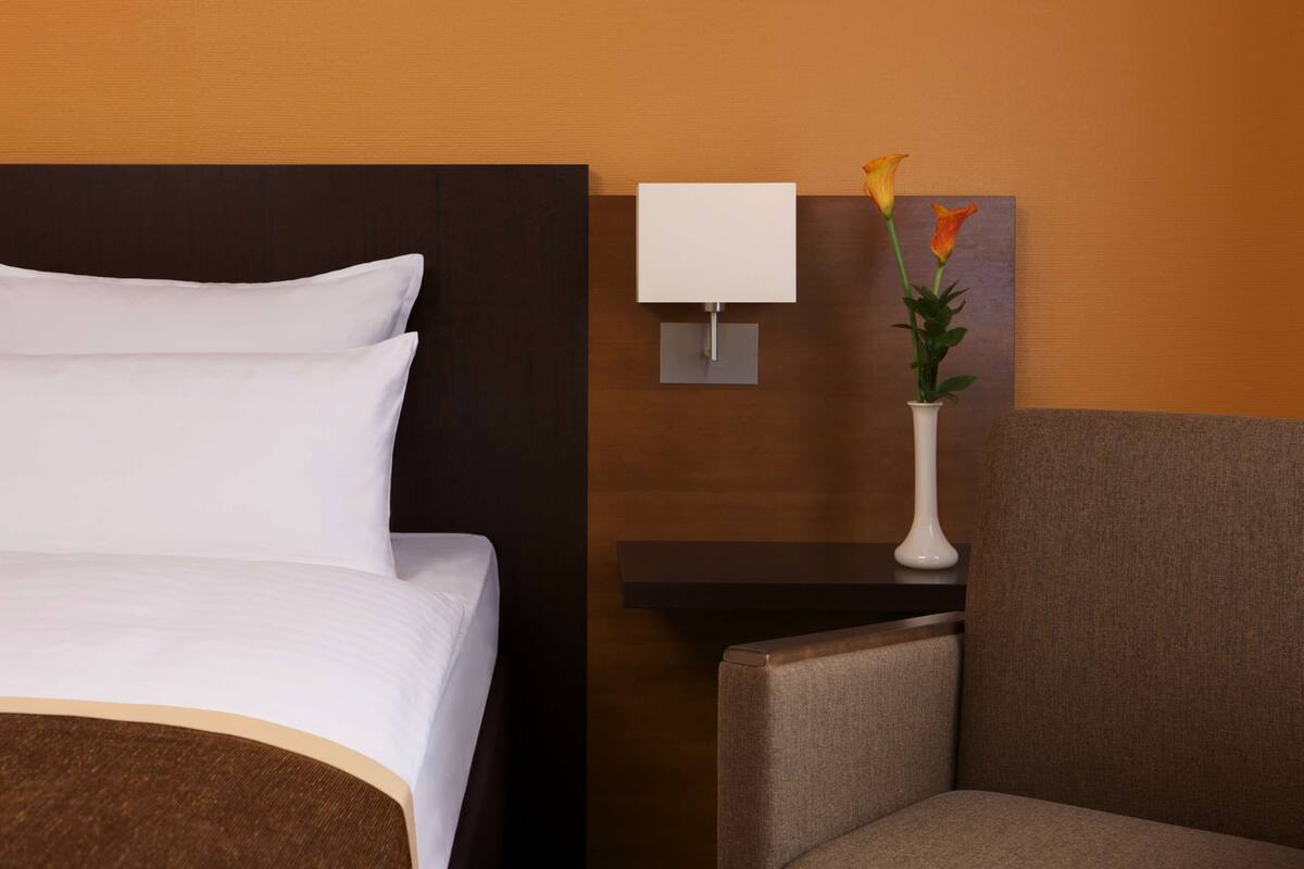 A hotel room with a bed, a nightstand with a lamp, a vase with flowers, and a chair.