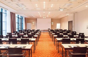 Un salón de conferencias con filas de mesas y sillas, una pantalla de proyector y un gran ventanal con cortinas.