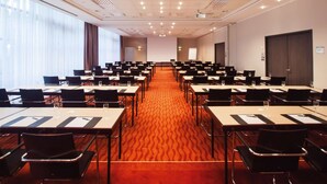 A conference room with rows of tables and chairs, a projector screen, and a large window with curtains. - Steigenberger Hotel Dortmund (Dortmund)
