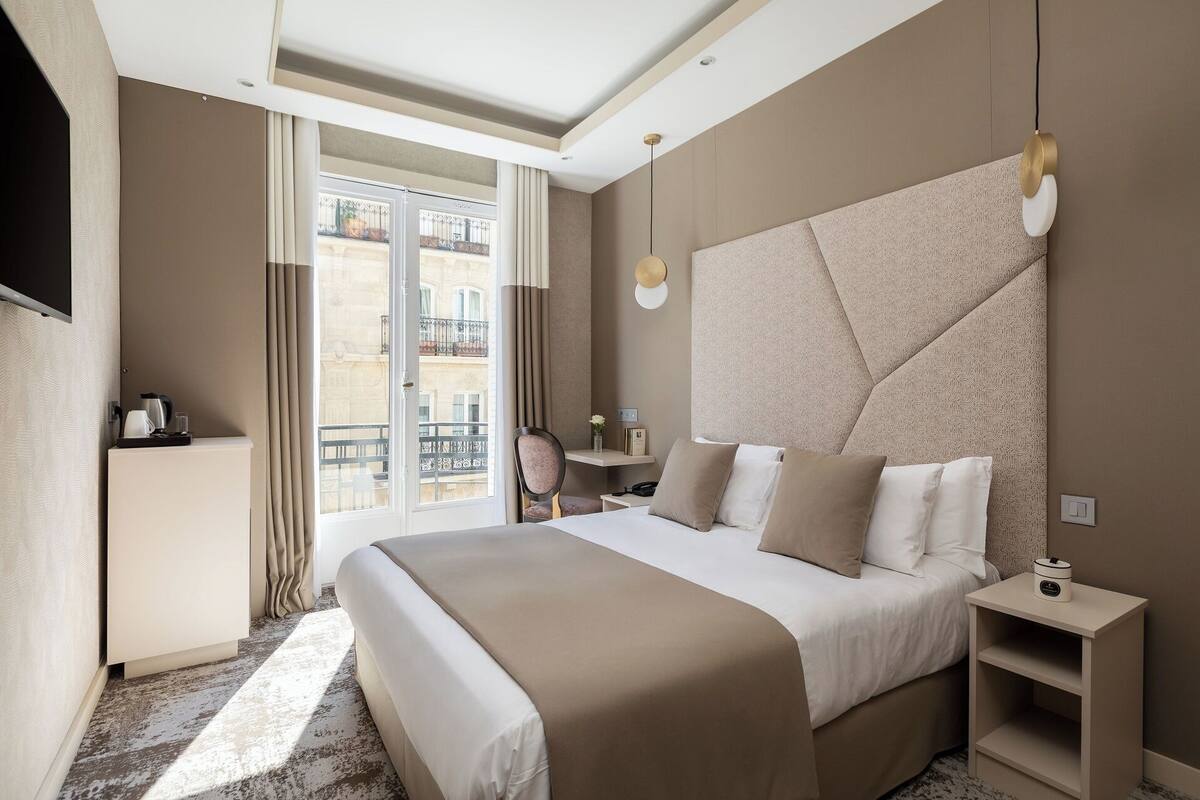 A modern hotel room with a large bed, a bedside table, a chair, and a view of the city through a large window.