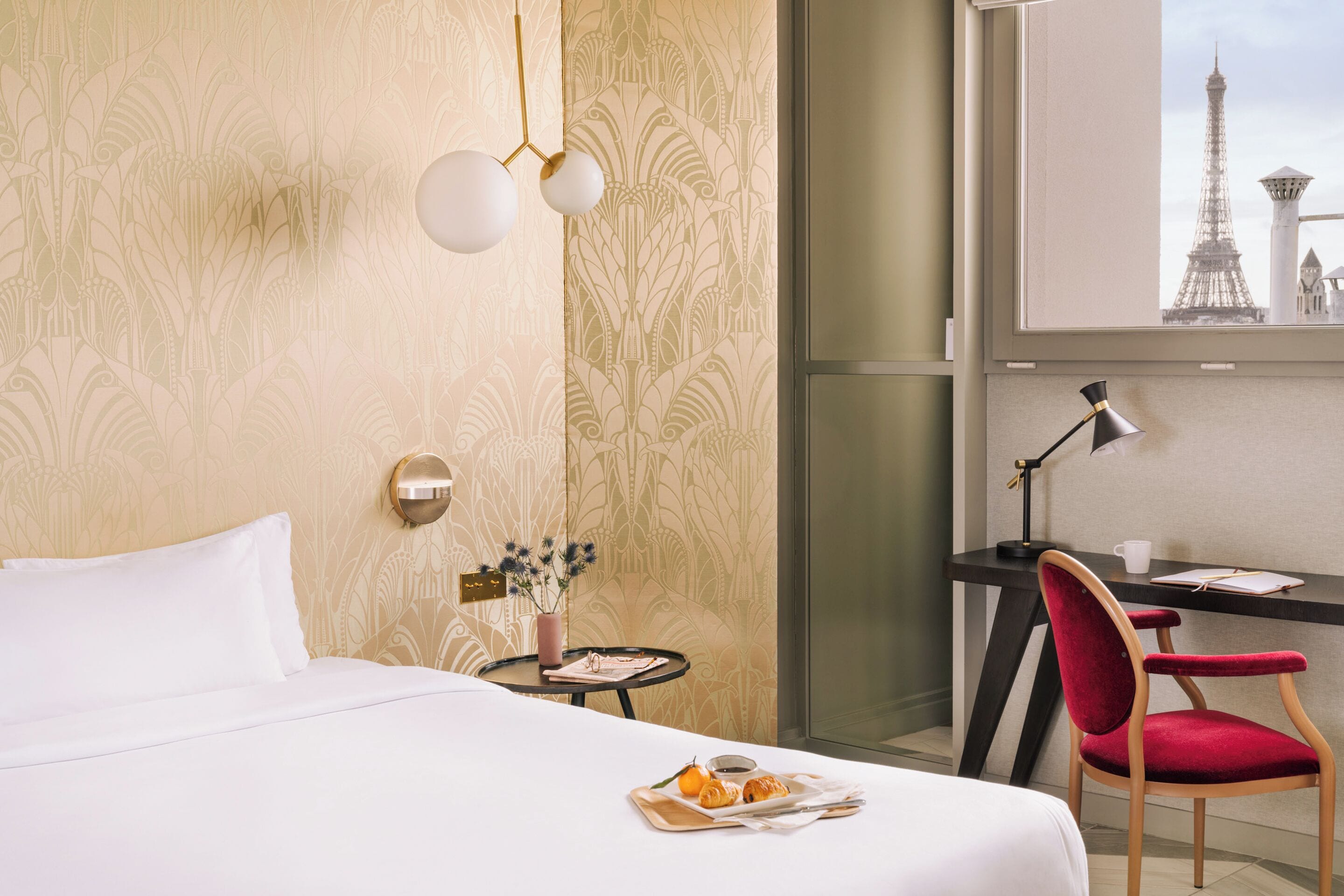A hotel room with a bed, a desk with a chair, a lamp, a small table with flowers, and a tray with pastries.