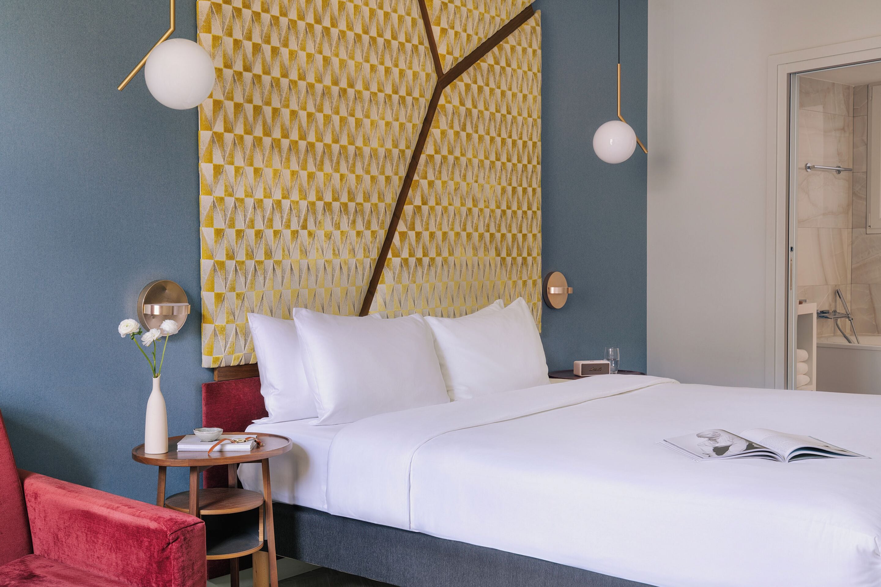 Ein Hotelzimmer mit einem Bett, einem roten Sessel, einem kleinen Tisch mit Blumenvase und einer Wand mit Muster.