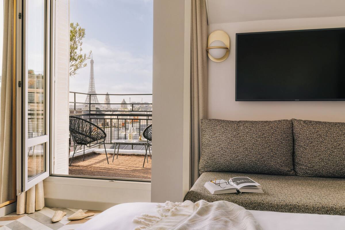 A hotel room with a bed, a sofa, a TV, and a balcony with a view of the Eiffel Tower.