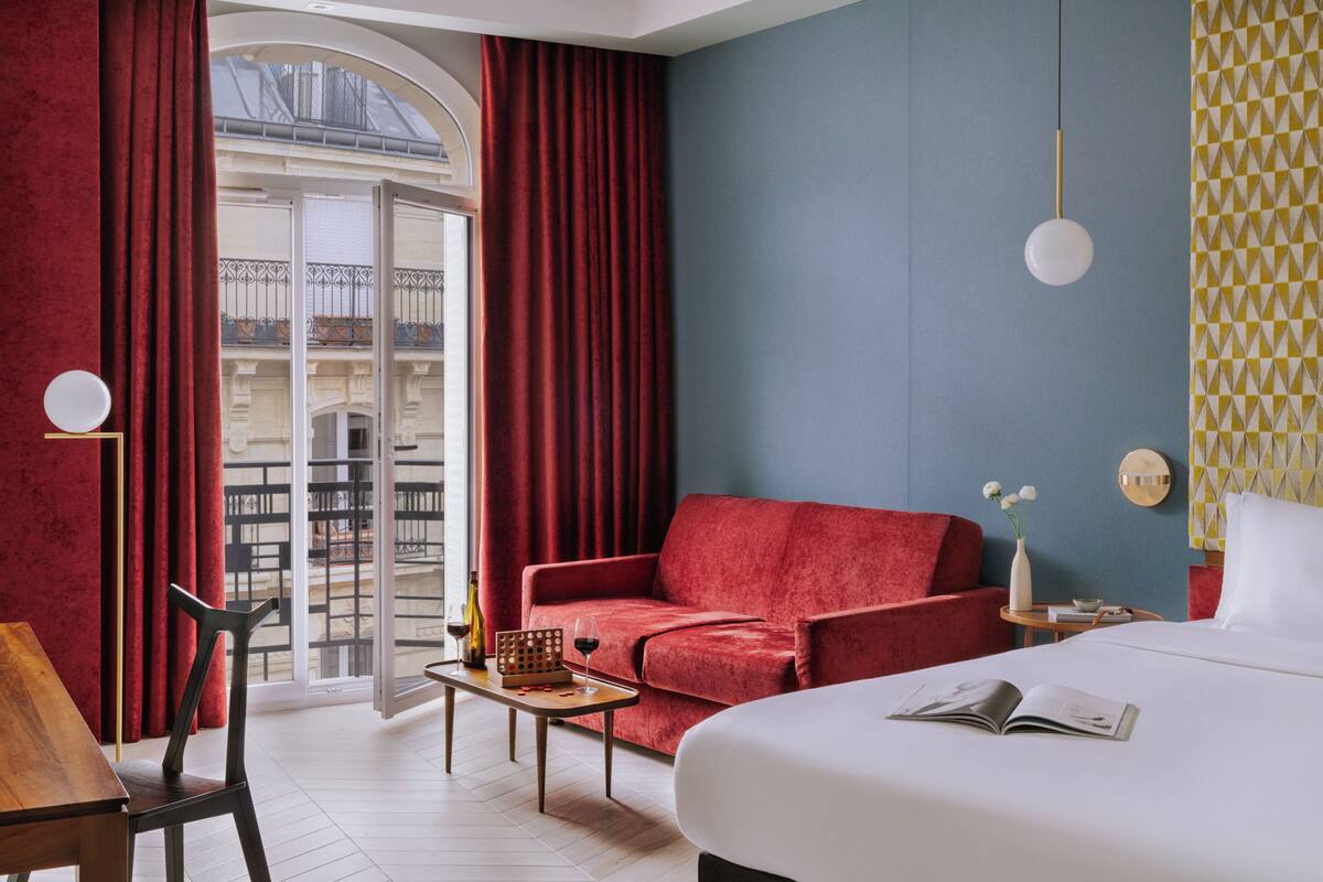 A hotel room with a red sofa, a bed, a wooden table, and a balcony with red curtains.