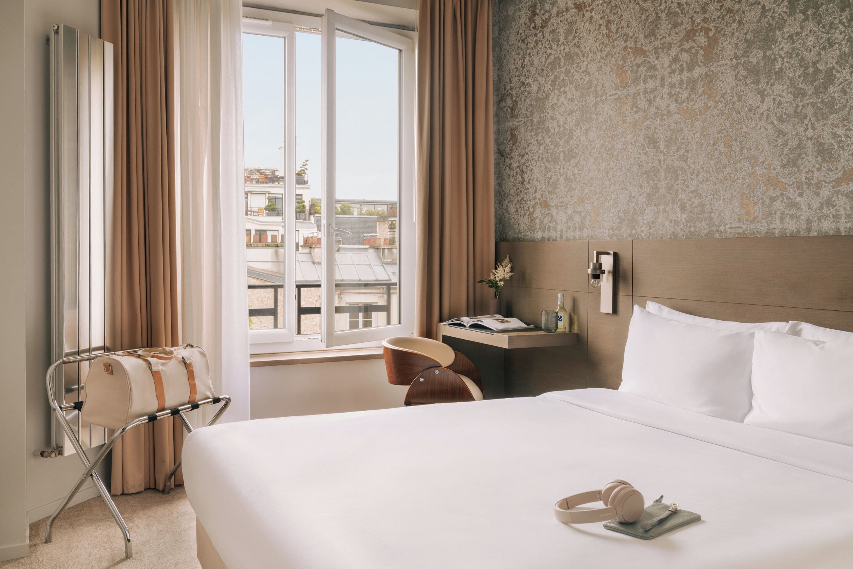 Une chambre d’hôtel avec un lit, une fenêtre avec des rideaux, une table de chevet avec une lampe, et une vue sur le paysage urbain.