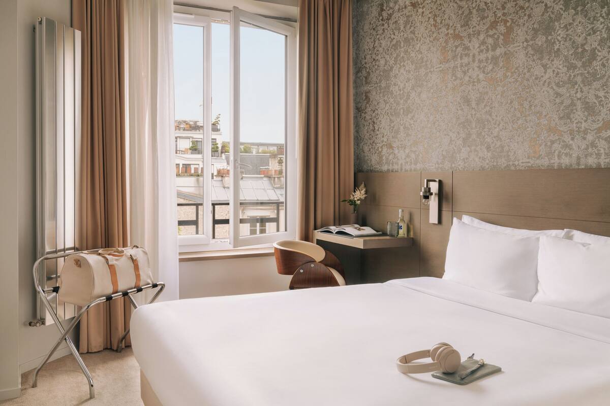 A hotel room with a bed, a window with curtains, a bedside table with a lamp, and a view of the cityscape.