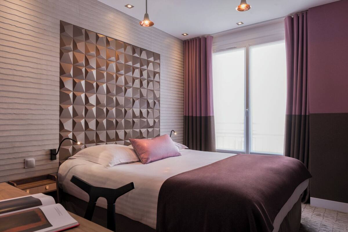 A modern hotel room with a geometric patterned headboard, a large bed with a dark-colored bedspread, a bedside table with a book, and a view of the outdoors through a large window with curtains.