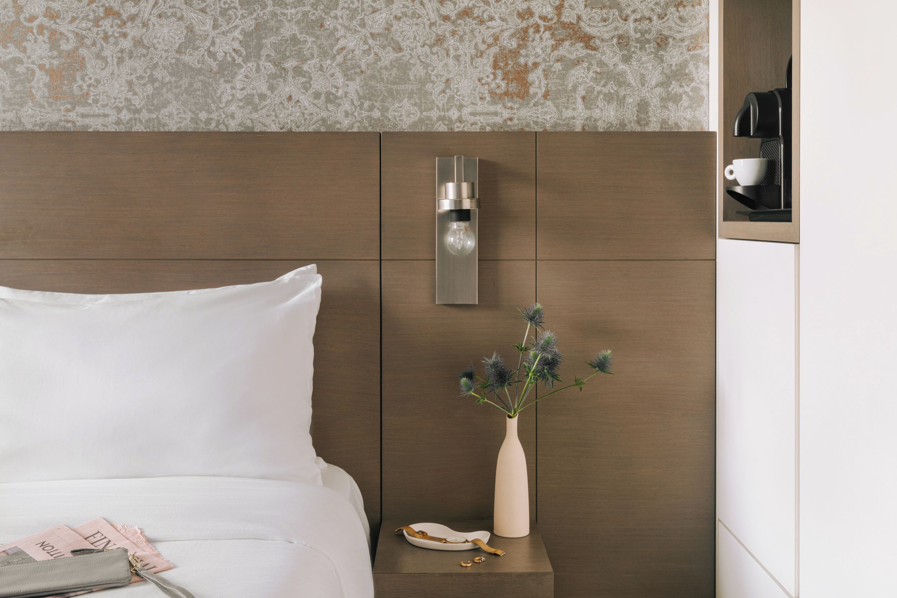 A hotel room with a bed, a nightstand with a vase of flowers, a coffee machine, and a wall-mounted light fixture.