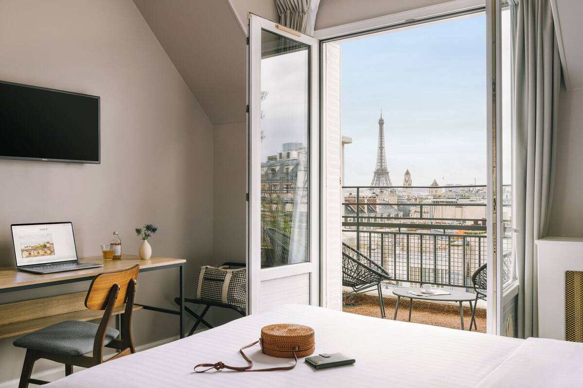 A hotel room with a balcony, a desk with a laptop, a bed, and a view of the Eiffel Tower.
