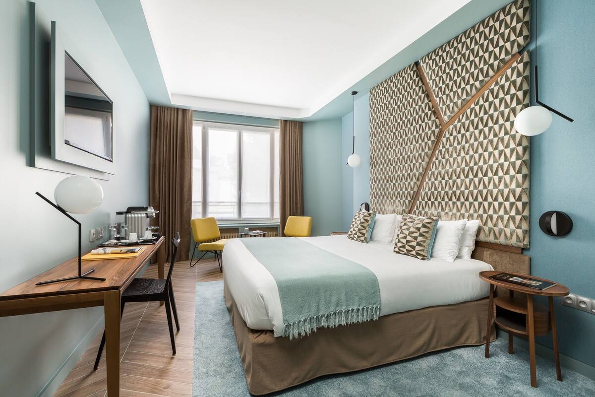 A modern hotel room with a large bed, a desk, a chair, and a window with curtains.