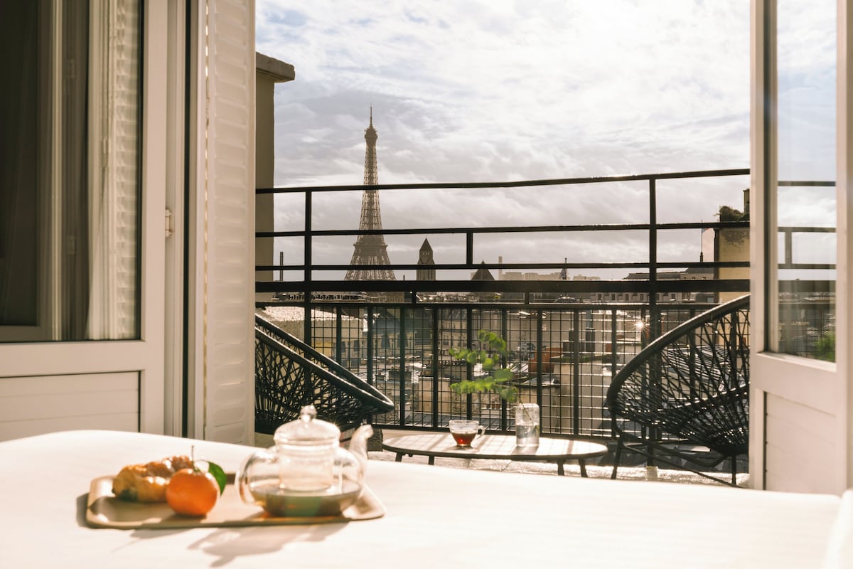 A balcony with a view of the Eiffel Tower, a table with a teapot and pastries, and a chair.