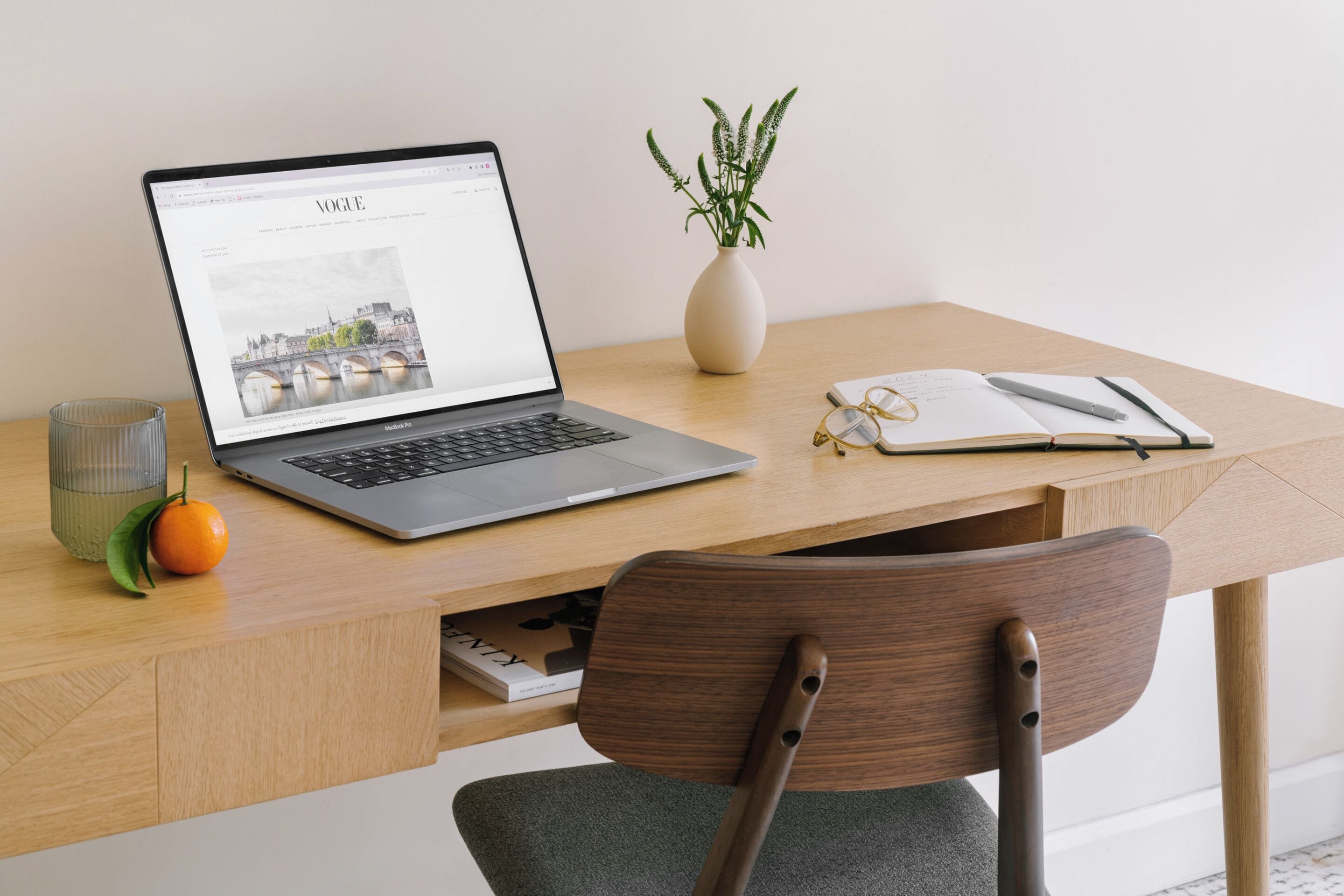 A laptop on a wooden desk with a notebook, pen, glasses, and a plant.