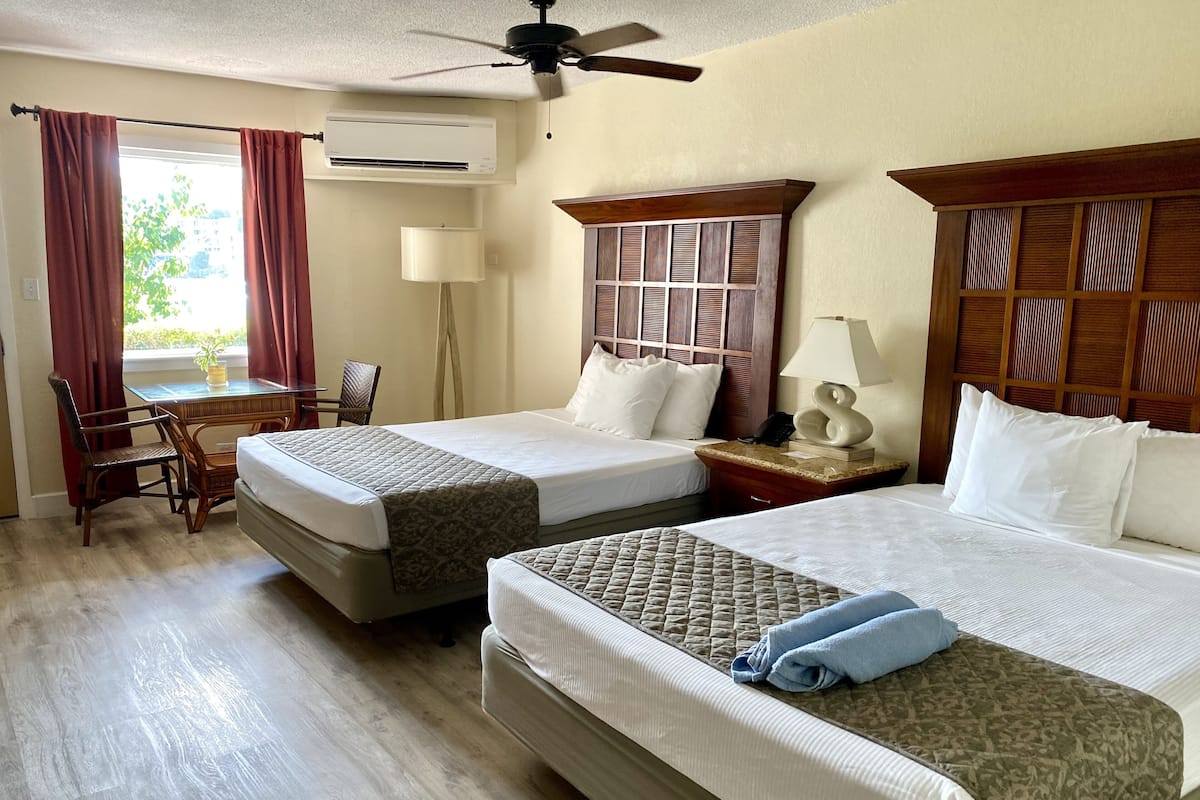 A hotel room with two beds, a desk with a chair, a lamp, and a ceiling fan.