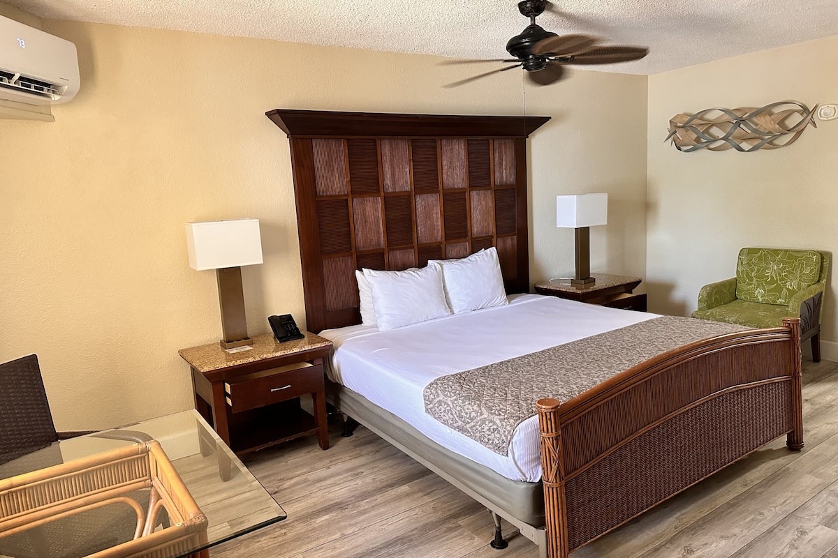 A hotel room with a large bed, a wooden headboard, a ceiling fan, a green armchair, a nightstand with a lamp, and a wicker chair.