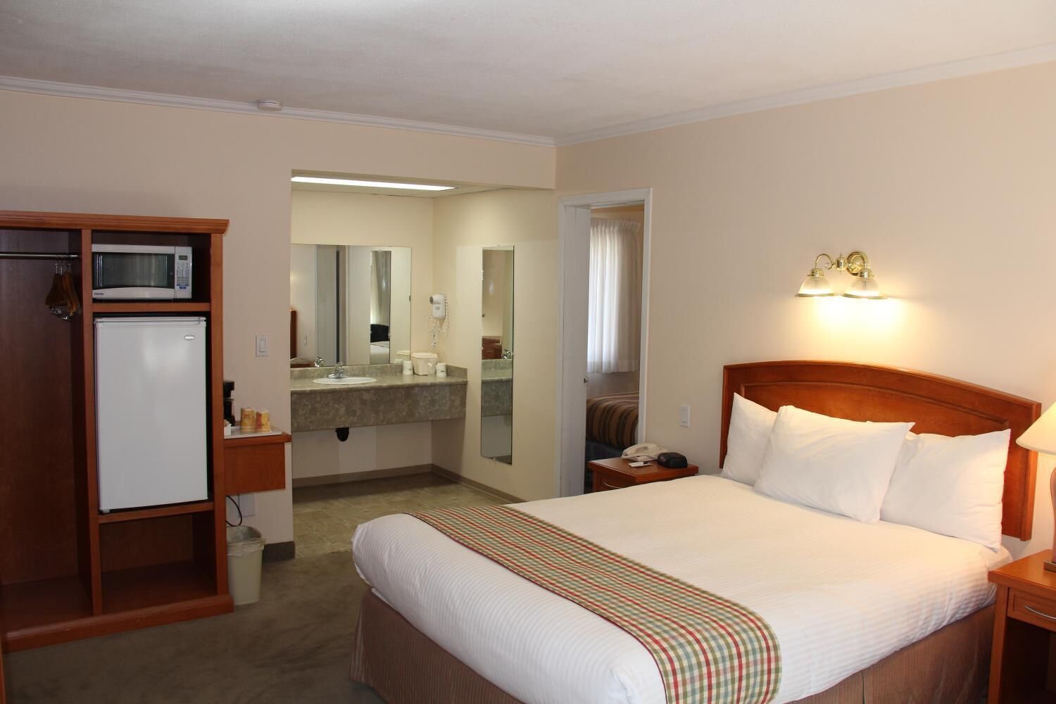 A hotel room with a bed, a dresser, a microwave, a refrigerator, a bathroom with a sink and mirror, and a nightstand with a lamp.