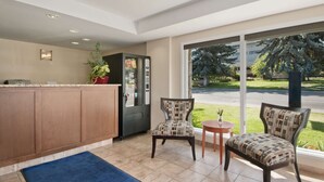 A reception area with a counter, two chairs, a small table, and a large window with a view of trees and a building. - Travelodge by Wyndham Kamloops (Kamloops)
