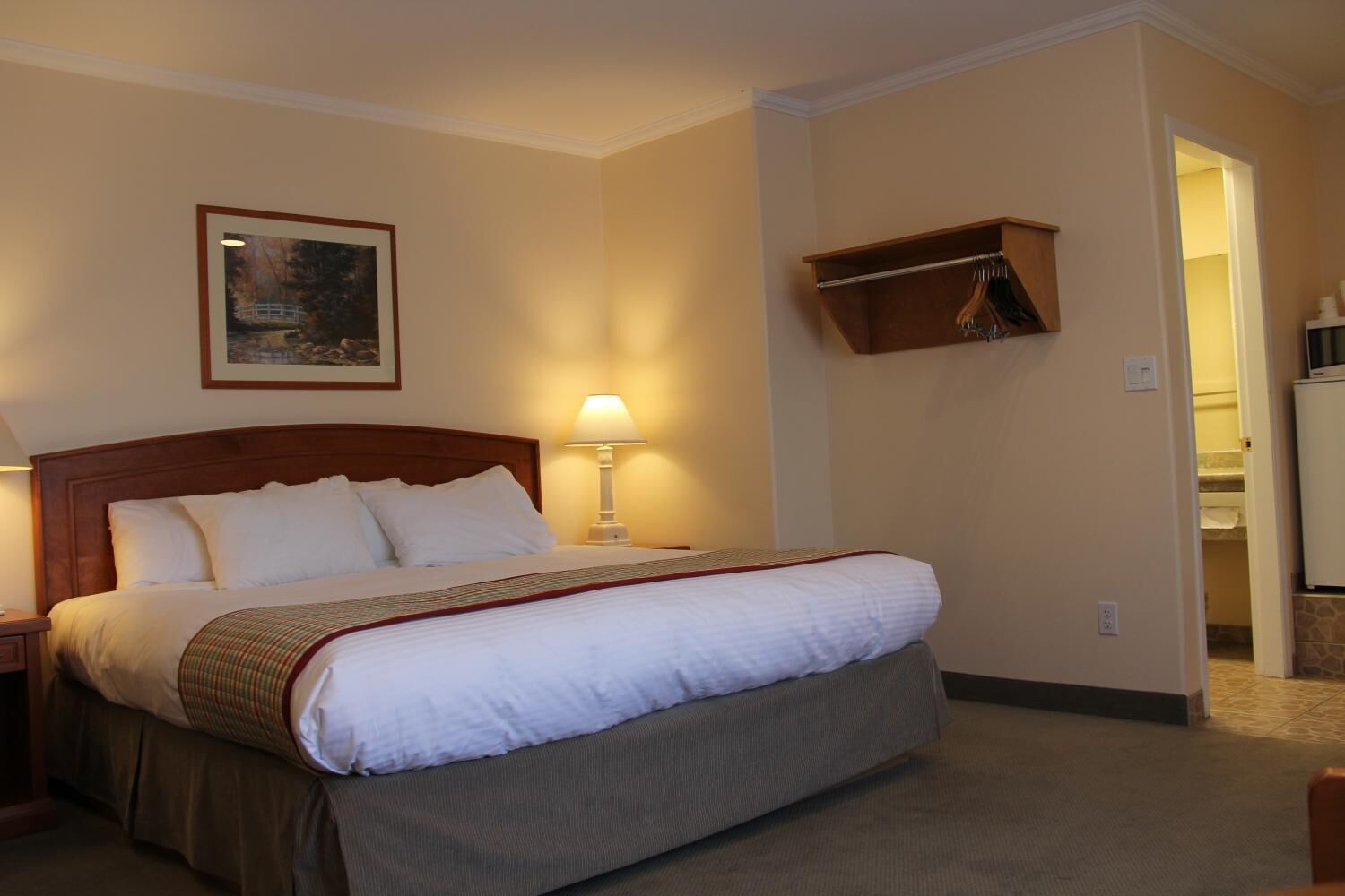 A hotel room with a large bed, a nightstand, a lamp, a framed picture on the wall, and a coat rack.