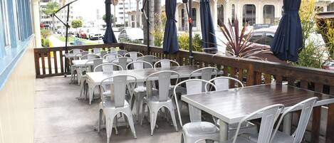 A balcony with white plastic chairs and tables, umbrellas, and a view of palm trees and buildings.