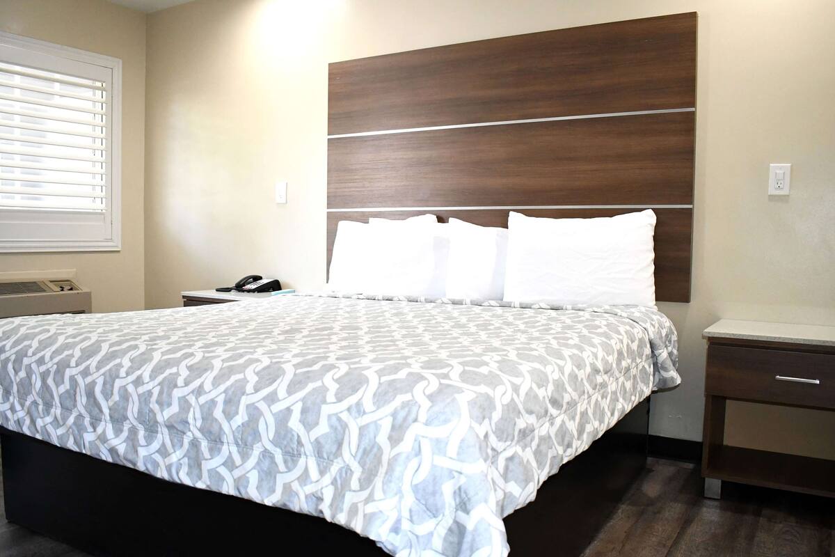 Een net opgemaakt bed met een bedspreide met patroon, een houten hoofdbord, een nachtkastje met lamp en een raam met jaloezieën.