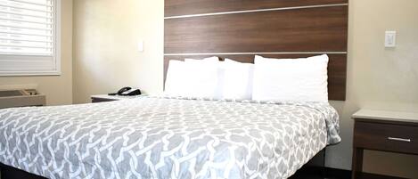 Una cama bien hecha con una colcha estampada, un cabecero de madera, una mesita de noche con una lámpara y una ventana con persianas.