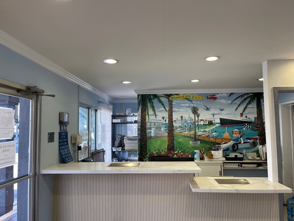 Zona de recepción con un mural de una escena de playa, un mostrador de recepción y una ventana con vistas al exterior.