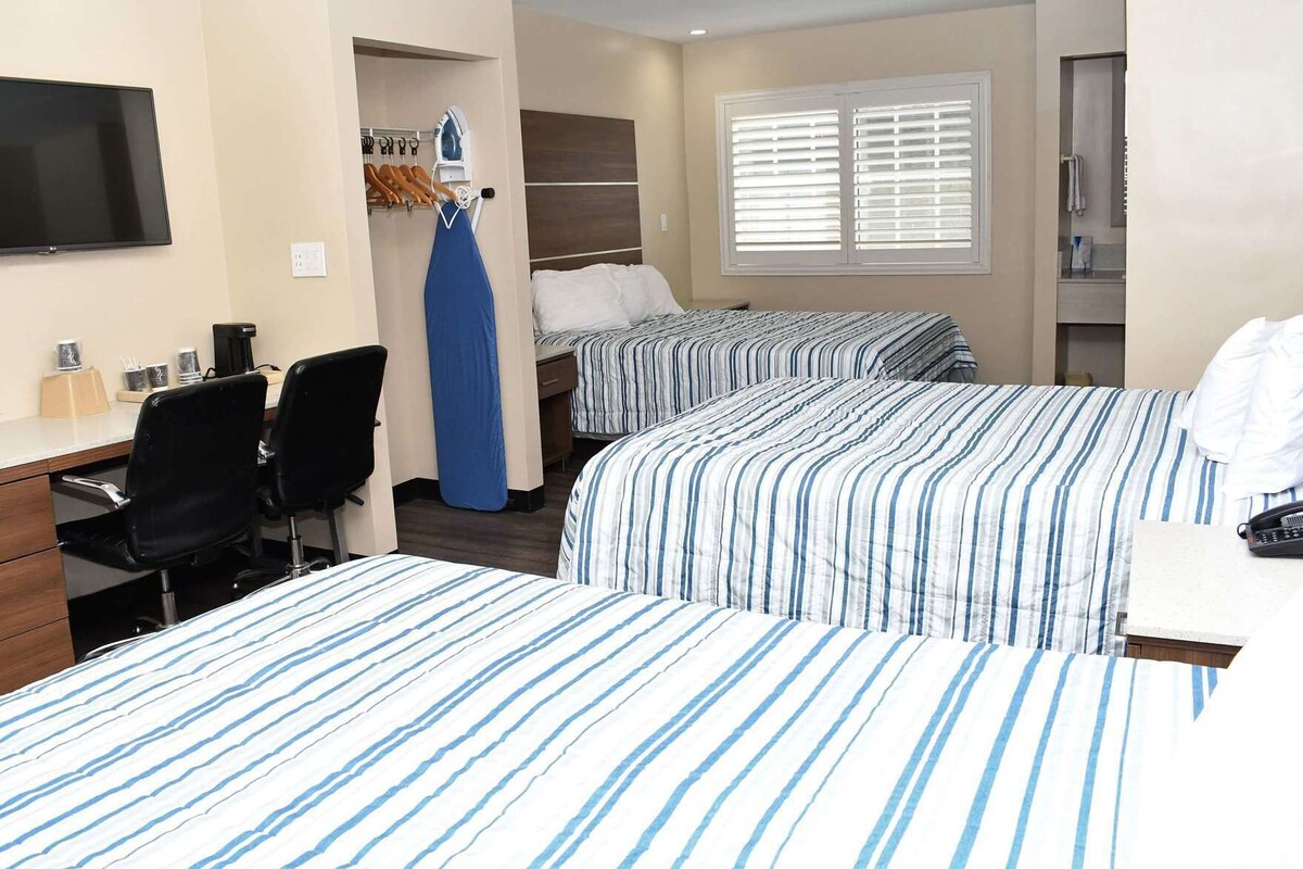Habitación de hotel con dos camas, escritorio, sillas, televisor y ventana con persianas.
