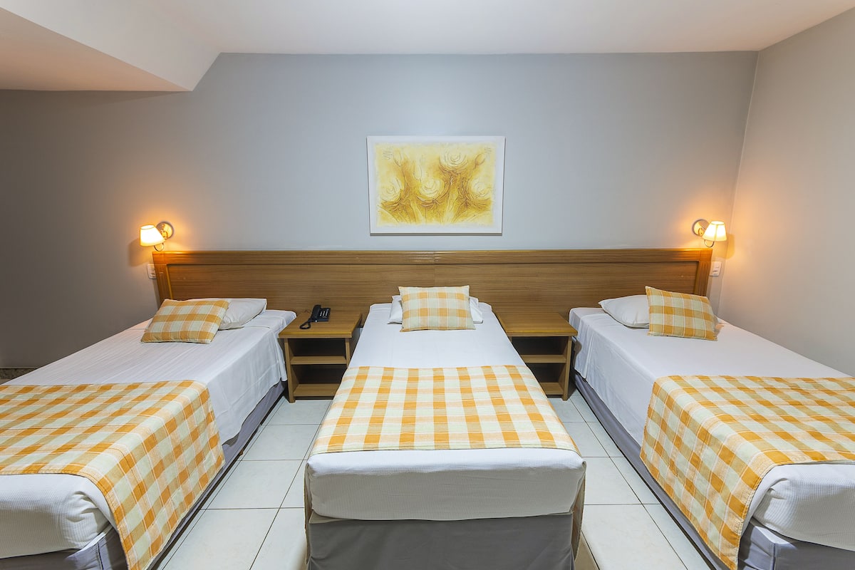 Habitación de hotel con tres camas individuales, cada una con una colcha de cuadros y almohadas a juego. Cuenta con un cabecero de madera, una obra de arte en la pared y lámparas de noche.